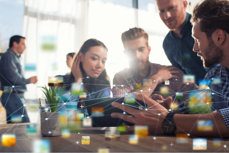 A group of people collaborates on a digital project at a desk surrounded by floating icons and network connections.