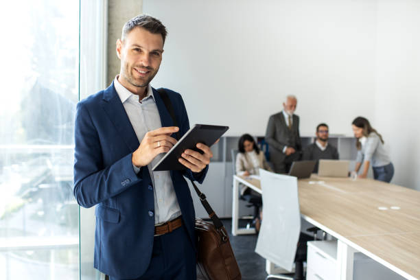 Man in a suit using a tablet in a modern office, with colleagues working in the background.