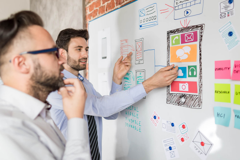 Two men are discussing and pointing at a large design sketch on a whiteboard full of diagrams and sticky notes.