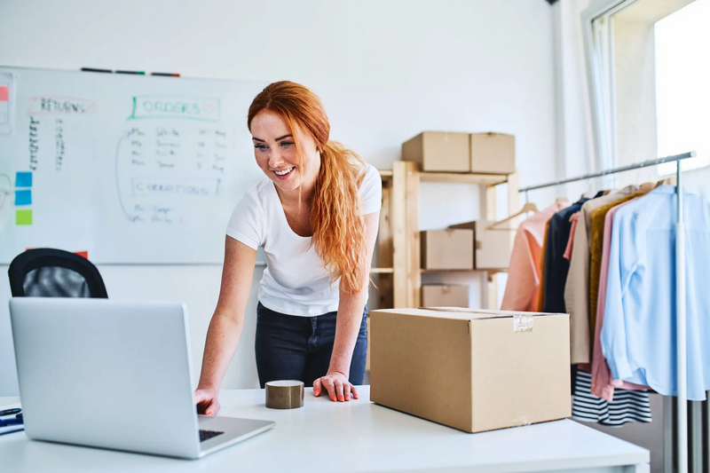 A woman works at a desk with a laptop, a cardboard box, and clothes hanging in the background.