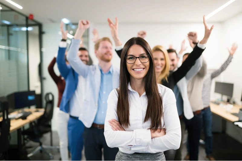 A woman smiles with her arms crossed while a group of people behind her raise their hands in celebration in an office setting.