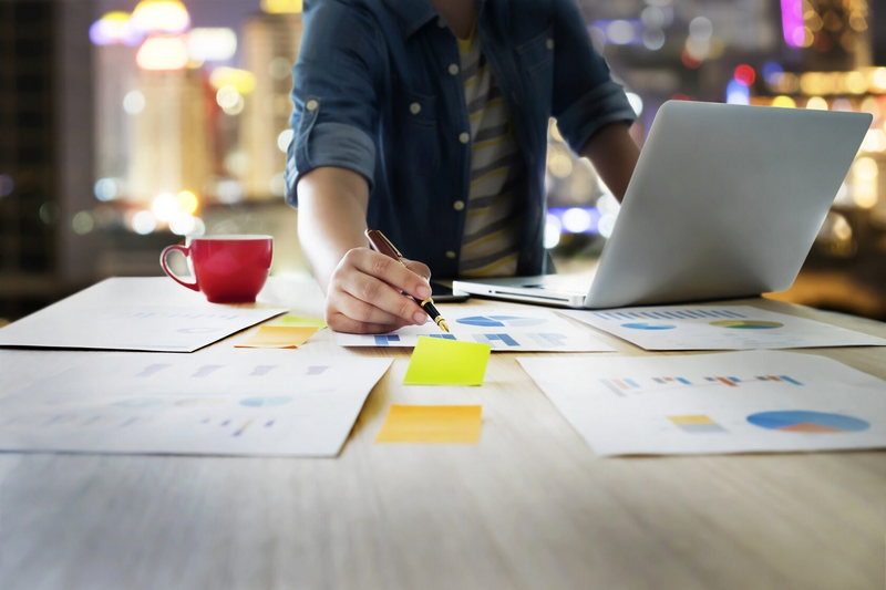 Person working on a laptop at a desk covered with charts and sticky notes, with a cup of coffee nearby.