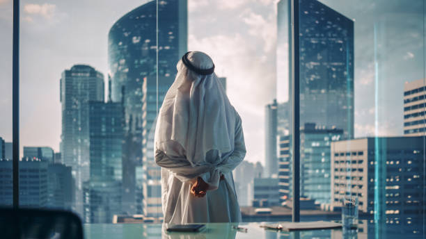 A person in traditional attire stands in a modern office overlooking the skyscrapers of a UAE city.