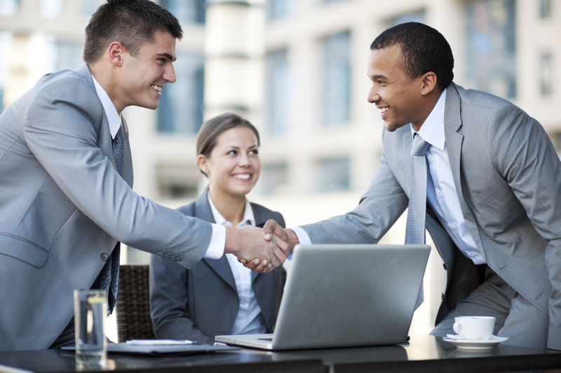 Two businessmen in suits shake hands while a businesswoman looks on, all seated around a laptop.