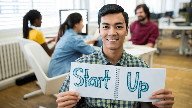 A smiling man in an office holds a notebook with Start Up written on it, while colleagues work in the background.